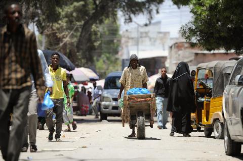 Tur privat de 3 zile în Mogadiscio (istorie, cultură și coastă) A street scene in Mogadishu features people walking, a man pulling a cart, a woman in a niqab, and various vehicles including a tuk-tuk.