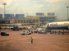 This image shows a busy airport tarmac with planes, vehicles, and people, in front of a building labeled "Aéroport de Ndjili".