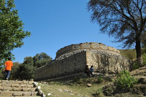 A man ascends stone steps toward a large, round, stone structure with two people sitting at its base, under a clear blue sky.