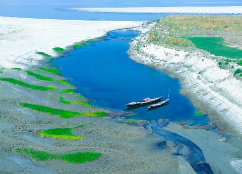 An aerial view of a tranquil blue river with two boats, surrounded by sandy shores and patches of green vegetation.