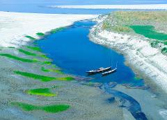 An aerial view of a tranquil blue river with two boats, surrounded by sandy shores and patches of green vegetation.