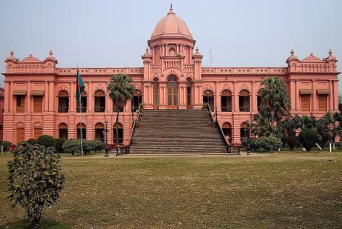 The pink Ahsan Manzil palace with a grand staircase, a dome, and a Bangladesh flag stands prominently in a grassy field.