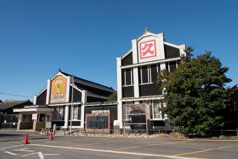 A traditional black wooden building with a large red symbol on its gable stands under a clear blue sky, hinting at the historical significance of miso production.
