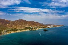 Aerial view of a scenic coastline with brown mountains, a sandy beach, and turquoise water, punctuated by a small rocky island and boats, under a blue sky with scattered clouds.