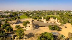An aerial shot showcases the Al Jahili Fort in Al Ain, UAE, with its distinctive circular tower, surrounded by walls, trees, and the urban landscape in the background.
