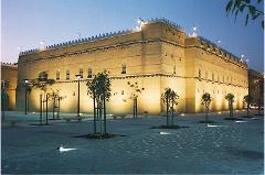 A beige fort-like building stands illuminated at night, with trees and benches in the plaza, creating an atmospheric urban scene.