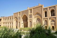A large, beige building with arched doorways and intricate designs, partially obscured by tall green grasses, stands under a clear blue sky.
