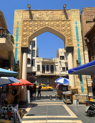 A bustling marketplace street is framed by an ornate, archway made of pale bricks, with Islamic calligraphy and geometric patterns.