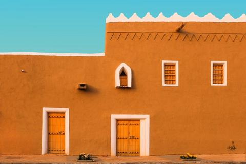 A sand-colored adobe building with white trim features two doors, two windows, and a small arched niche under a bright blue sky.