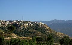 Eine Stadt liegt auf einem Hügel mit einer Felswand, im Hintergrund sind Berge und im Vordergrund Laub zu sehen.
