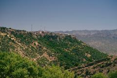 A scenic view of a town perched atop a cliff overlooking a lush, green hillside with mountains in the distance under a clear blue sky.