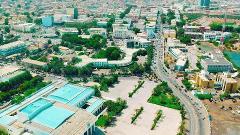 An aerial shot of a cityscape featuring buildings, trees, a wide street with traffic, and a large open area with palm trees.
