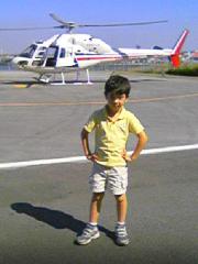 Un niño posa con confianza, con las manos en las caderas, frente a un helicóptero blanco y rojo en un aeródromo.