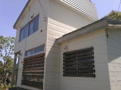 A light-colored, modern house with horizontal siding and slatted windows is surrounded by trees and a clear blue sky.