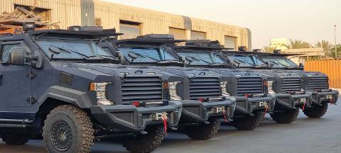 A line of dark gray armored vehicles stands parked in front of a building with corrugated metal siding.