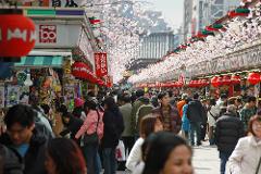 A bustling crowd moves through a market street lined with shops adorned with red lanterns and cherry blossoms.