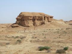 A large, weathered sandstone formation with cave-like openings sits in a dry, arid landscape with sparse vegetation under a clear sky.