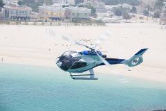 A helicopter flies over the turquoise water near a sandy beach with buildings in the background.