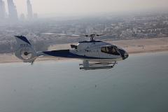 A blue and white helicopter flies over the sea, with a city skyline in the background.