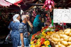 Day Tour in Bazurto Market At an outdoor market, shoppers browse a colorful display of produce, including peppers, potatoes, and tomatoes, under red awnings, with a sign indicating the price of chili peppers.
