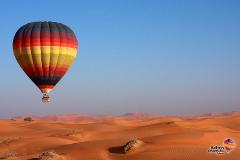 A hot air balloon floats over a vast, sandy desert landscape under a clear blue sky.
