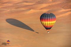 A hot air balloon floats over a vast, undulating desert landscape, casting a long shadow on the sand.