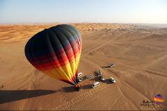 A hot air balloon hovers above a desert landscape with several vehicles waiting below.