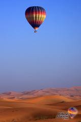 A colorful hot air balloon soars above a desert landscape with rolling sand dunes under a clear blue sky, with the "Balloon Adventures Emirates" logo in the bottom right.