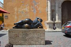 Gran Tour incluyendo Bogotá, Medellín, Nuquí y Valle del Cocora - 11 Días A Botero sculpture of a reclining woman rests on a brick pedestal against a weathered wall, near a car and an old building in a sunny outdoor setting.