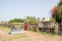 A sign for the Badagry Heritage Museum sits in front of a weathered fence and gate, with trees and a building visible in the background.