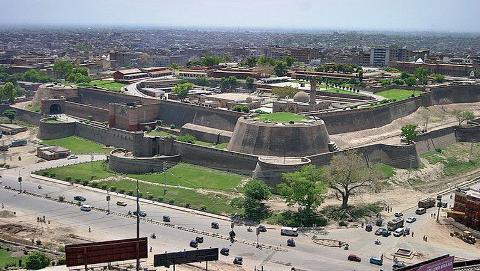An aerial shot reveals the Lahore Fort, surrounded by walls, greenery, and a bustling cityscape with cars and buildings.