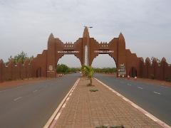 This eye-level shot showcases the Welcome to Bamako gate, a large mud-brick structure spanning a paved road, with "Welcome to Bamako" in English and French, flanked by matching walls, under an overcast sky.