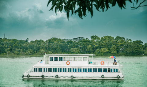 A white boat sits on calm, greenish water with lush green trees lining the shore, beneath an overcast sky and some leafy branches in the foreground.