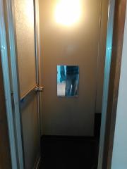 A person's legs are reflected in a small square mirror on a beige wall in a narrow hallway.