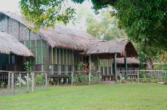 A picturesque wildlife lodge with thatched roofs and wooden stilts stands amidst lush greenery and trees, surrounded by a fence, on the banks of the Bensbach River in Far West PNG.