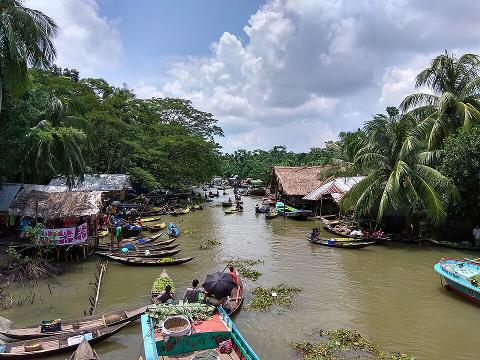 A vibrant scene unfolds at a Bangladeshi floating market, where numerous boats gather, laden with goods and vendors, along a river lined with stilted houses and lush greenery.