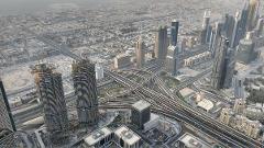An aerial view of Dubai, UAE, showing a large freeway interchange and a cluster of high-rise buildings on a slightly hazy day.