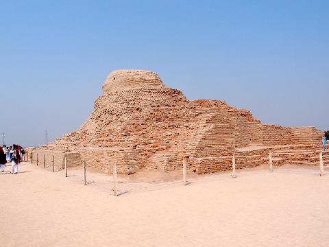 A large, ancient brick structure stands under a clear blue sky, surrounded by a sandy plain and a simple rope barrier.