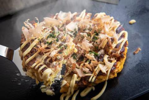 A close-up shot captures a steaming slice of okonomiyaki being lifted by a metal spatula, revealing layers of cabbage, meat, and a generous drizzle of mayonnaise and bonito flakes.