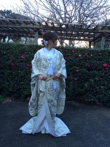A woman in a traditional white Japanese kimono stands in front of a flower bush under a wooden trellis, looking off to the side.