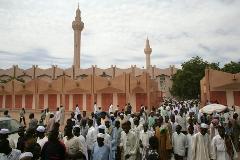 A large crowd of people is gathering in front of a mosque with two tall minarets under a cloudy sky.