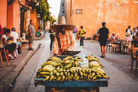 Day Tour in Bazurto Market A man with a large straw hat stands behind a cart full of plantains on a street in a colorful city, with people sitting and walking in the background.