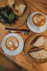 Coffeeholic Tour in Medellin A table displaying a light meal, featuring two cups of artful lattes, a sandwich cut in half, and toasted bread with a side salad.