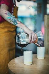 Coffeeholic Tour in Medellin A person with a colorful tattoo on their arm is pouring coffee from a Chemex into two white cups on a wooden table.