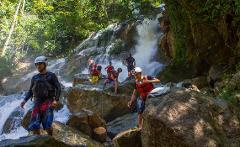 A group of people wearing helmets and life vests navigates a rocky waterfall in a lush, green environment.