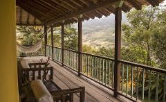 This is a cozy porch with a wooden railing, wooden furniture, and a hammock, offering a scenic view of the surrounding lush greenery and distant hills.