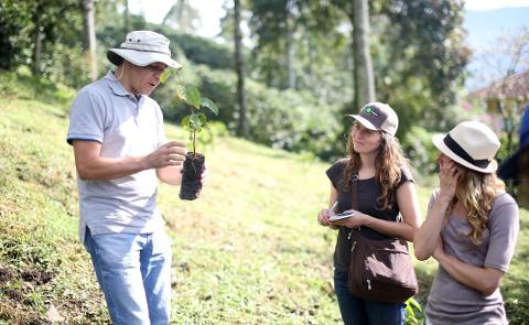 A man in a hat shows a small plant in a black bag to two women, one of whom is writing in a notebook.