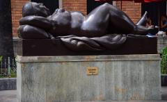 A bronze sculpture of a reclining woman rests on a marble base in a public space, with people visible in the background.