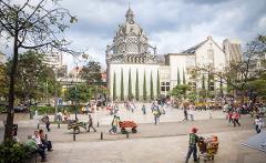 A bustling city square in Medellín, Colombia, with the Metropolitan Cathedral as the focal point, surrounded by people, trees, and vendor carts under a cloudy sky.