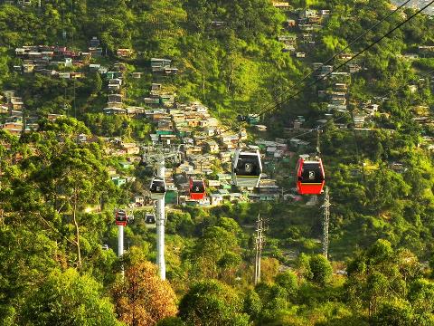 A hillside covered in houses is traversed by a cable car system, with red and white gondolas traveling above lush green trees.